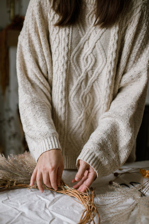Hands holding dried grass, herbs, leaves, ribbon and scissors on rustic table. Woman making stylish autumn wreath. Fall decor and arrangement in home.の写真素材