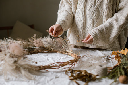 Woman making stylish autumn wreath. Hands with dried grass, herbs, leaves, ribbon and scissors on rustic table. Details of fall decor and arrangement in homeの写真素材