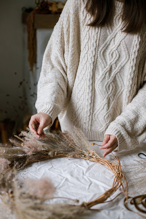 Woman making stylish autumn wreath. Hands with dried grass, herbs, leaves, ribbon and scissors on rustic table. Details of fall decor and arrangement in homeの写真素材