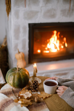 Hands in cozy sweater holding warm cup of tea with pumpkin and autumn leaves on table on background of burning fireplace. Autumn hygge. Thanksgiving and Halloween.の写真素材