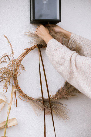 Woman decorating house entrance with stylish autumn wreath. Rustic wreath with dried grass, herbs and ribbon in hands. Fall decor of front door and arrangement. Thanksgiving and Halloweenの写真素材