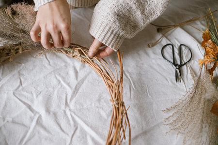 Hands making stylish autumn wreath. Dried grass, herbs, leaves, ribbon and scissors on rustic table. Details of fall decor and arrangement in home. Thanksgiving and Halloweenの写真素材