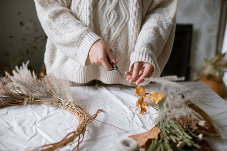 Hands making stylish autumn wreath. Dried grass, herbs, leaves, ribbon and scissors on rustic table. Details of fall decor and arrangement in home. Thanksgiving and Halloweenの写真素材