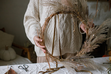 Hands holding wreath with dried grass, herbs, leaves, ribbon on rustic table. Woman making stylish autumn wreath. Fall decor and arrangement in home.の写真素材