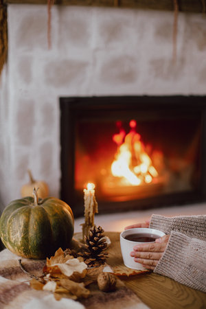 Autumn hygge. Hands holding cup of tea and pumpkin, autumn leaves on table on background of warm fireplace. Cozy fall in living room. Thanksgiving and Halloween.の写真素材