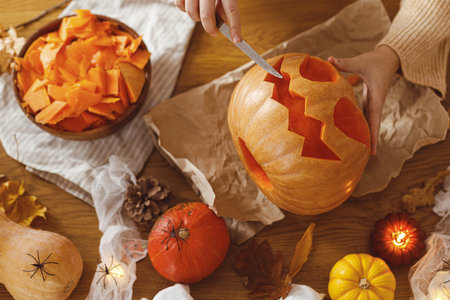 Making jack o lantern. Woman carving halloween pumpkin on table with ghosts and spiders, halloween preparation. Happy halloween! Making festive decorationsの写真素材
