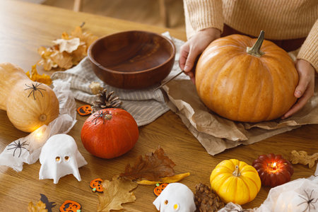 Woman holding pumpkin and knife on table with ghosts and spiders in living room. Carve a pumpkin, halloween preparation. Making halloween decorations, space for text.の写真素材