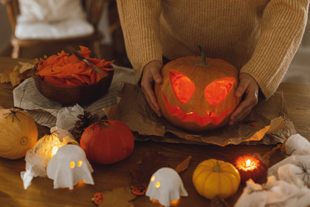 Carving pumpkin. Woman holding glowing carved halloween pumpkin on table with ghosts and spiders. Making jack o lantern. Happy halloween!の写真素材
