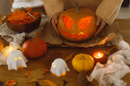 Making jack o lantern. Woman holding glowing carved halloween pumpkin on table with ghosts and spiders, halloween preparation. Happy halloween! Making festive decorationsの写真素材