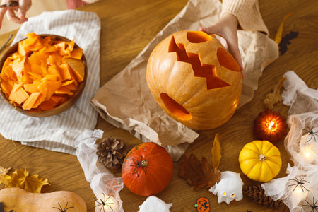 Woman carving halloween pumpkin on table with ghosts and spiders. Carving jack o lantern, halloween preparation. Making halloween decorationsの写真素材
