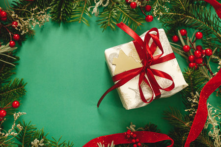 Stylish Christmas gift flat lay. Modern christmas present with red ribbon and green fir branches, red berries and gold ornaments on green background. Xmas banner. Season's greeting cardの写真素材