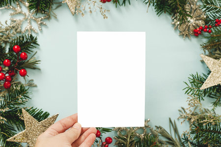 Christmas card mock up. Hand holding empty greeting card on blue background with fir branches, red berries and golden ornaments. Merry Christmas. Season greeting postcard templateの写真素材