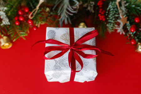 Merry Christmas! Stylish Christmas gift with red ribbon and green fir branches, red berries and gold ornaments on red background. Xmas background. Season's greetingsの写真素材