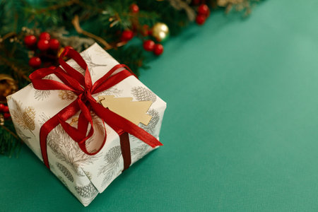 Stylish Christmas gift with red ribbon and green fir branches, red berries and gold ornaments on green background. Season's greetings, space for text. Merry Christmas!の写真素材