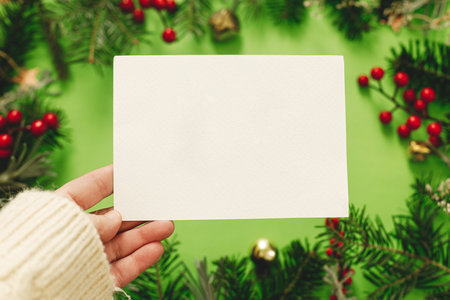 Christmas card mock up. Hand holding empty greeting card on green background with fir branches, red berries and golden ornaments. Merry Christmas. Season greeting postcard templateの写真素材