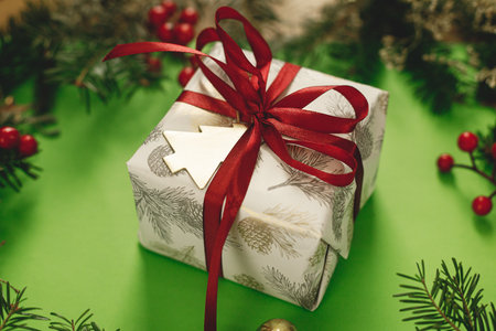Merry Christmas! Stylish Christmas gift with red ribbon and green fir branches, red berries and gold ornaments on green background. Xmas background. Season's greetingsの写真素材