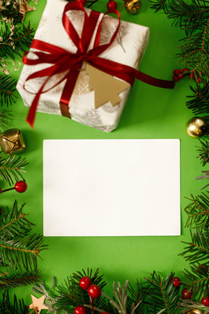Christmas card mock up. Empty greeting card flat lay with fir branches, red berries and gift box on green background. Merry Christmas. Season greetings templateの写真素材