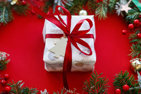 Stylish Christmas gift. Modern christmas present with red ribbon and green fir branches, red berries  on red background. Xmas background. Season's greeting cardの写真素材