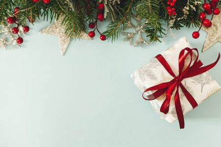 Merry Christmas! Stylish Christmas gift with red ribbon and fir branches, red berries and glitter ornaments on blue background flat lay. Season's greetings, space for text.の写真素材