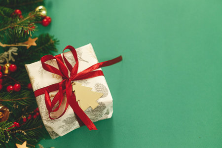 Stylish Christmas gift with red ribbon and green fir branches, red berries and gold ornaments on green background flat lay. Season's greetings, space for text. Merry Christmas!の写真素材