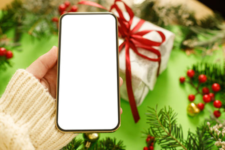 Hand holding smartphone with empty screen on green background with fir branches, red berries and gift box. Christmas phone mock up. Space for text. Christmas advertising, app templateの写真素材