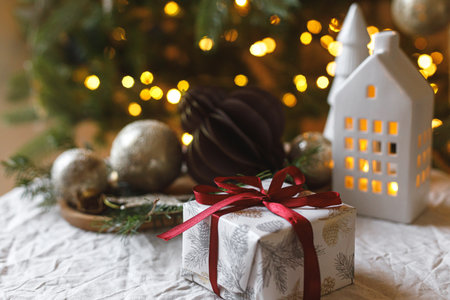 Merry Christmas! Stylish Christmas gift with red ribbon, baubles and decor with fir branches on rustic table on background of christmas tree lights. Winter holiday aestheticの写真素材