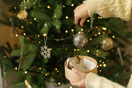 Decorating christmas tree with vintage golden baubles. Hands in cozy sweater holding stylish Christmas ornaments on background of christmas tree with golden lights. Merry Christmasの写真素材
