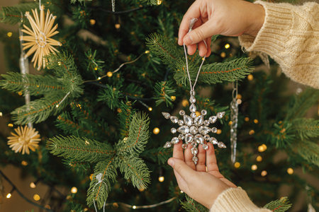 Hands in knitted sweater hanging stylish Christmas ornament on christmas tree with golden lights. Decorating christmas tree with vintage glass snowflakeの写真素材