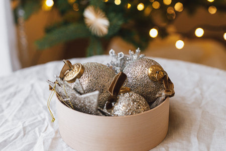 Decorating christmas tree with vintage golden baubles. Stylish Christmas ornaments in box on rustic table on background of christmas tree with lights. Merry Christmasの写真素材