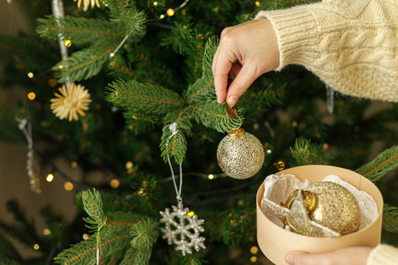 Hands in knitted sweater holding stylish Christmas ornaments in wooden box on background of christmas tree with golden lights. Decorating christmas tree with vintage golden baublesの写真素材