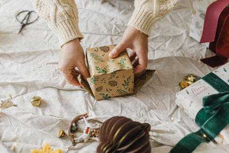 Hands wrapping christmas gift box with festive paper on table with linen cloth and ornaments, close up. Merry Christmas and Happy Holidaysの写真素材