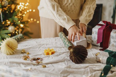 Wrapping stylish christmas gift, winter holidays preparation. Woman in cozy sweater holding gift box and festive paper on rustic table on background of decorated christmas tree with lightsの写真素材