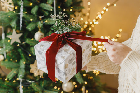 Merry Christmas and Happy Holidays. Hands in sweater opening modern christmas gift box with red ribbon and fir branch on background of stylish christmas tree with lights.の写真素材
