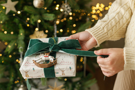 Merry Christmas and Happy Holidays. Hands in sweater opening modern christmas gift box with green ribbon and vintage toy on background of stylish christmas tree with lights.の写真素材