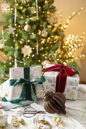 Stylish christmas gifts with spruce branch and modern ornaments on background of decorated christmas tree with golden lights. Merry Christmas and Happy Holidays. Modern Christmas presentの写真素材