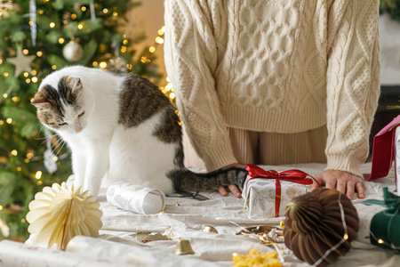 Wrapping stylish christmas gift, winter holidays preparation. Hands in cozy sweater decorating gift box and and cute cat helping on table on background of decorated christmas tree with lightsの写真素材