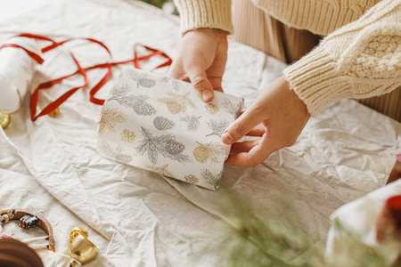Hands wrapping christmas gift box with festive paper on table with linen cloth and ornaments, close up. Merry Christmas and Happy Holidaysの写真素材