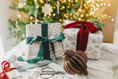 Merry Christmas and Happy Holidays. Stylish christmas gifts with spruce branch and modern ornaments on background of christmas tree golden lights. Christmas present holiday bannerの写真素材