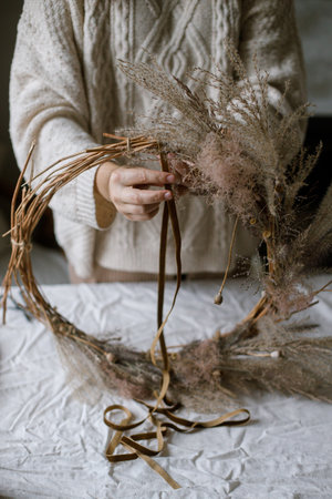 Woman making stylish autumn wreath. Hands holding wreath with dried grass, herbs, leaves, ribbon on rustic table. Details of fall decor and arrangement in homeの写真素材