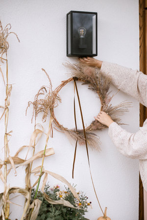 Woman decorating house entrance with stylish autumn wreath. Rustic wreath with dried grass, herbs and ribbon in hands. Fall decor of front door and arrangement. Thanksgiving and Halloweenの写真素材