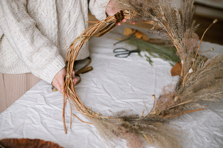 Hands making stylish autumn wreath. Dried grass, herbs, leaves, ribbon and scissors on rustic table. Details of fall decor and arrangement in home. Thanksgiving and Halloweenの写真素材