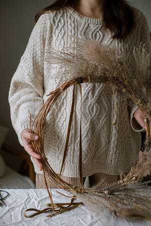 Woman making stylish autumn wreath. Hands holding wreath with dried grass, herbs, leaves, ribbon on rustic table. Details of fall decor and arrangement in homeの写真素材