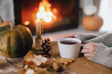 Autumn hygge. Hands holding cup of tea and pumpkin, autumn leaves on table on background of warm fireplace. Cozy fall in living room. Thanksgiving and Halloween.の写真素材