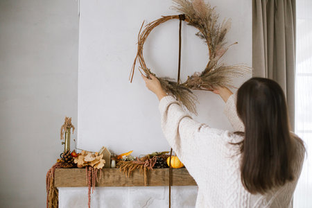 Woman decorating fireplace with stylish autumn wreath. Rustic wreath with dried grass, herbs and ribbon in hands. Fall decor and arrangement in home. Thanksgiving and Halloweenの写真素材