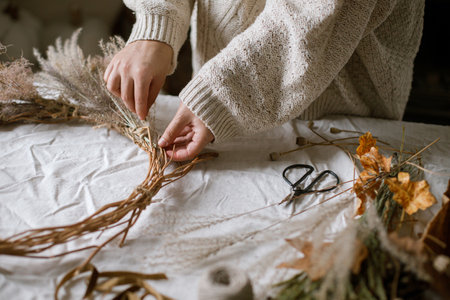 Hands making stylish autumn wreath. Dried grass, herbs, leaves, ribbon and scissors on rustic table. Details of fall decor and arrangement in home. Thanksgiving and Halloweenの写真素材