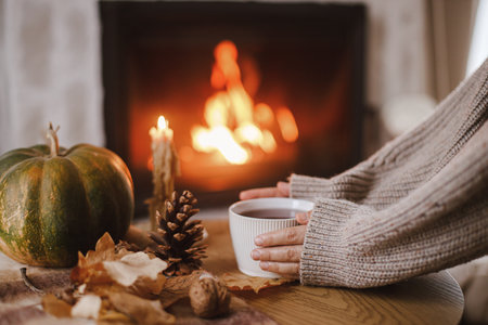 Hands in cozy sweater holding warm cup of tea with pumpkin and autumn leaves on table on background of burning fireplace. Autumn hygge. Thanksgiving and Halloween.の写真素材