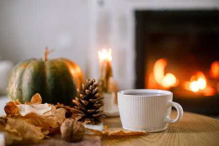 Cozy autumn. Cup of tea, pumpkin, autumn leaves on wooden table on background of warm fireplace. Fall still life in living room. Thanksgiving and Halloween.の写真素材