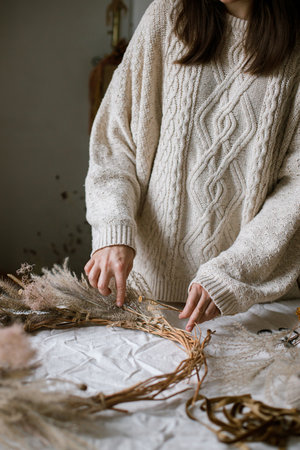 Hands holding dried grass, herbs, leaves, ribbon and scissors on rustic table. Woman making stylish autumn wreath. Fall decor and arrangement in home.の写真素材