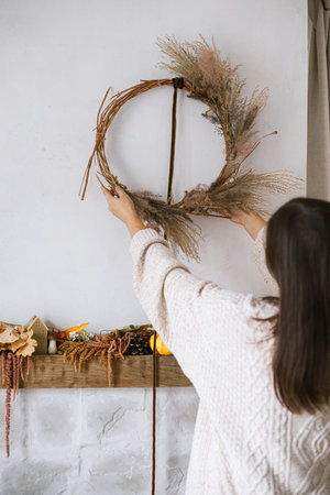 Woman decorating fireplace with stylish autumn wreath. Rustic wreath with dried grass, herbs and ribbon in hands. Fall decor and arrangement in home. Thanksgiving and Halloweenの写真素材
