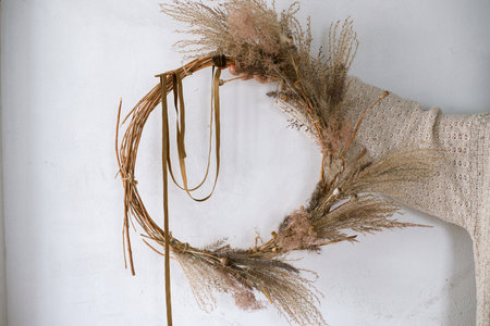Hands holding wreath with dried grass, herbs, leaves, ribbon at white wall. Woman making stylish autumn wreath. Fall decor and arrangement in home.の写真素材